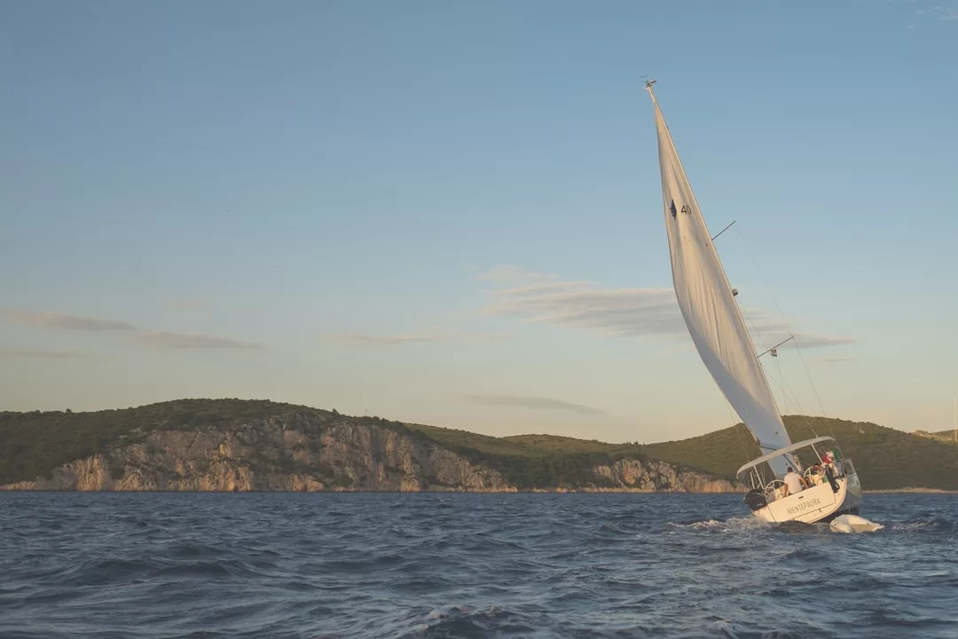 Un velero blanco navega sobre aguas azules agitadas cerca de la costa rocosa y arbolada de Mallorca, capturando la esencia del lujo bajo un cielo despejado.