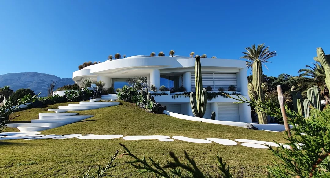 Casa moderna blanca con diseño curvo, rodeada de cactus y vegetación en un césped inclinado bajo el cielo azul claro de Mallorca, un verdadero ejemplo de vida de lujo.