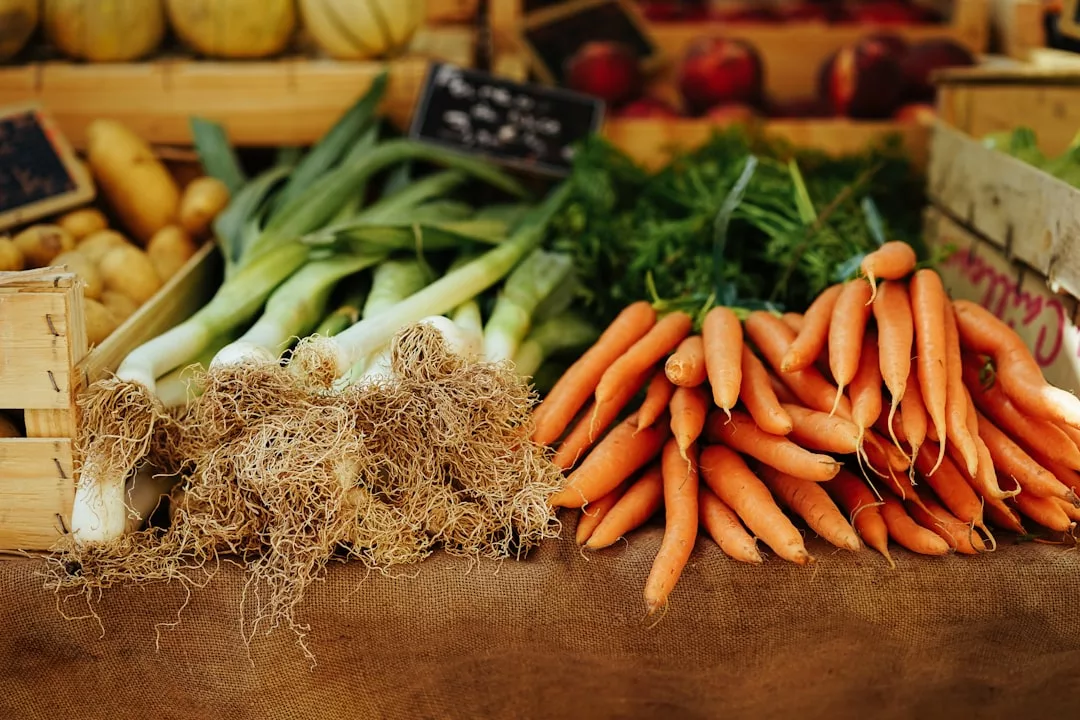 Puerros y zanahorias frescos expuestos en una mesa cubierta de arpillera en un mercado de Mallorca, con cajas de otros productos al fondo.