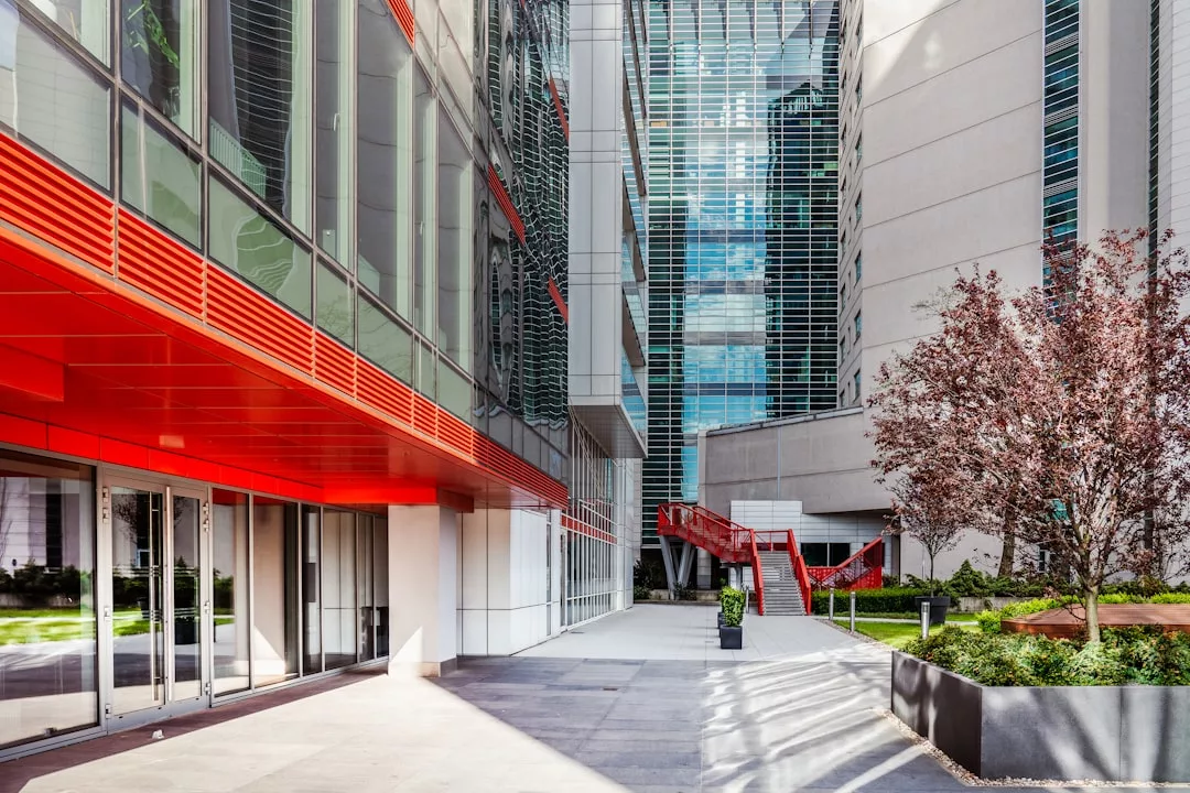 Moderno patio urbano con edificios de cristal, detalles arquitectónicos rojos, una escalera y jardineras con árboles y vegetación que evocan el relajado lujo de Mallorca.