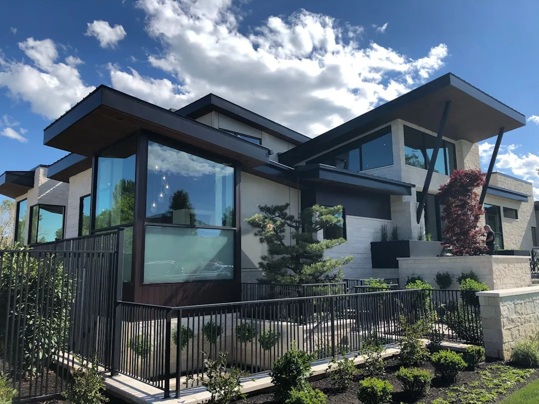 Moderna casa de dos plantas con grandes ventanales de cristal, tejado plano y patio delantero ajardinado bajo un cielo azul de Mallorca con nubes dispersas, perfecta para quienes buscan lujo y confort contemporáneo.