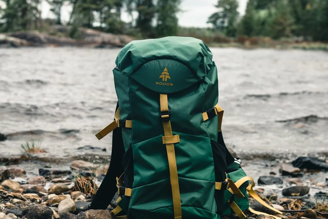 Una mochila verde y amarilla de la marca Woods descansa sobre una costa rocosa cerca de un río, evocando el relajado lujo de Mallorca, con árboles y agua visibles al fondo.