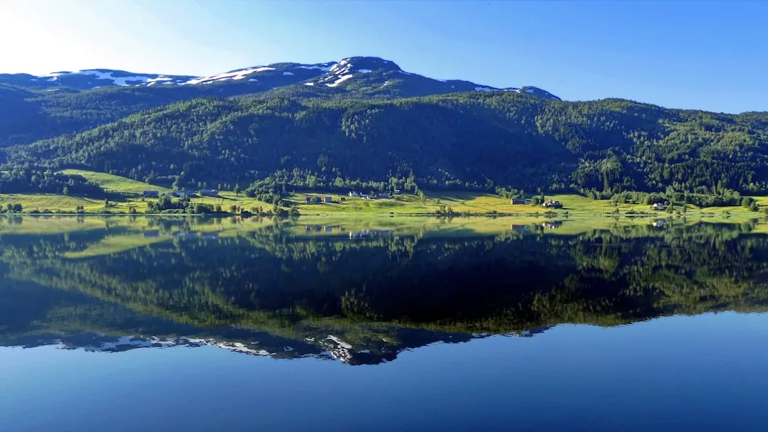 Un lago en calma refleja colinas verdes, casas dispersas y una montaña nevada bajo un cielo azul despejado, evocando el lujo y la tranquilidad de los serenos paisajes de Mallorca.