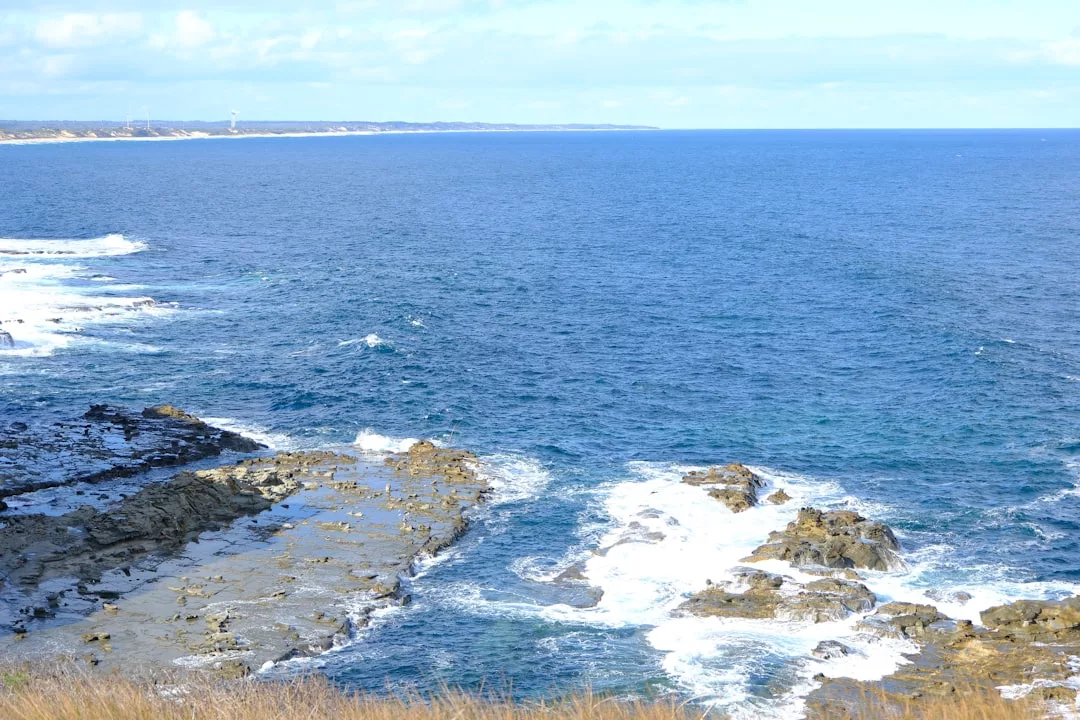 Costa rocosa con olas rompiendo contra las rocas bajo un cielo parcialmente nublado, con la pintoresca costa de Mallorca visible en la distancia - una verdadera escapada costera de lujo.