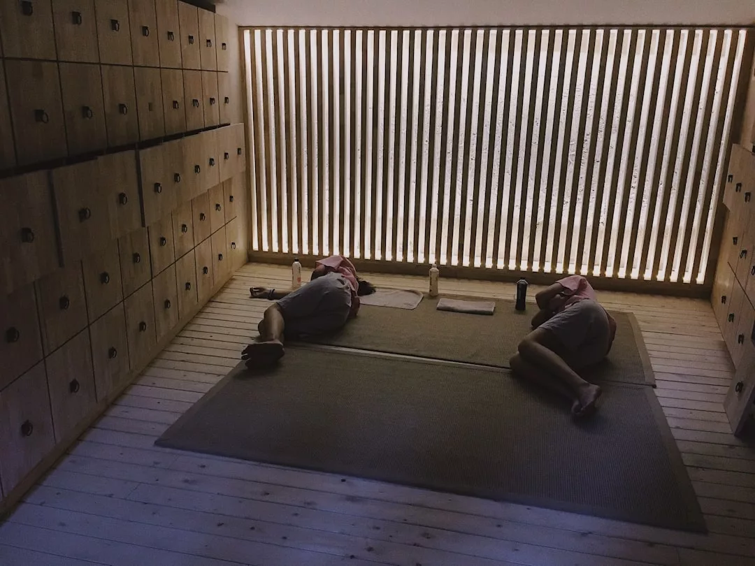 Dos personas están tumbadas sobre colchonetas en una habitación poco iluminada con taquillas de madera a un lado y listones verticales de madera en la otra pared, disfrutando de un momento de relajación de lujo que evoca la tranquilidad de Mallorca.