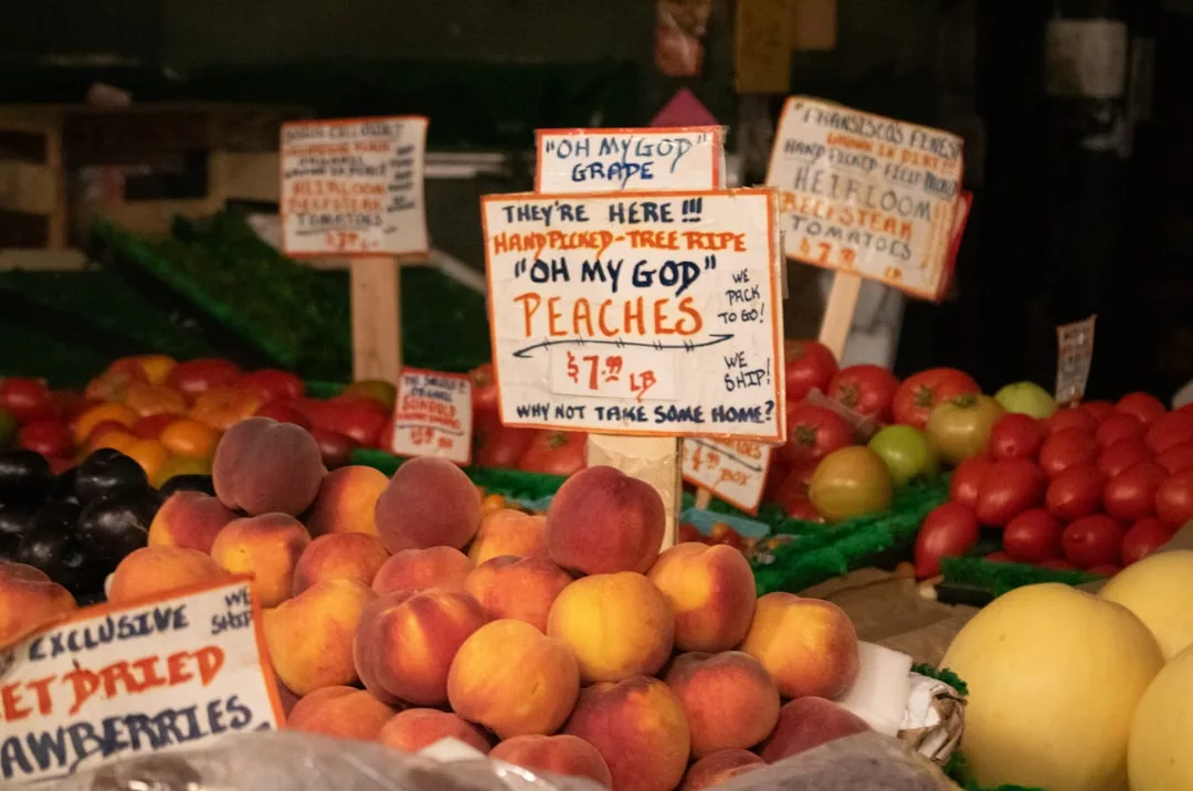 Un expositor de melocotones frescos a la venta en un mercado de Mallorca con un cartel escrito a mano que reza "Oh My God Peaches $7.99/lb" entre otras frutas y carteles de productos escritos a mano, aportando un toque de lujo a tu experiencia de compra.