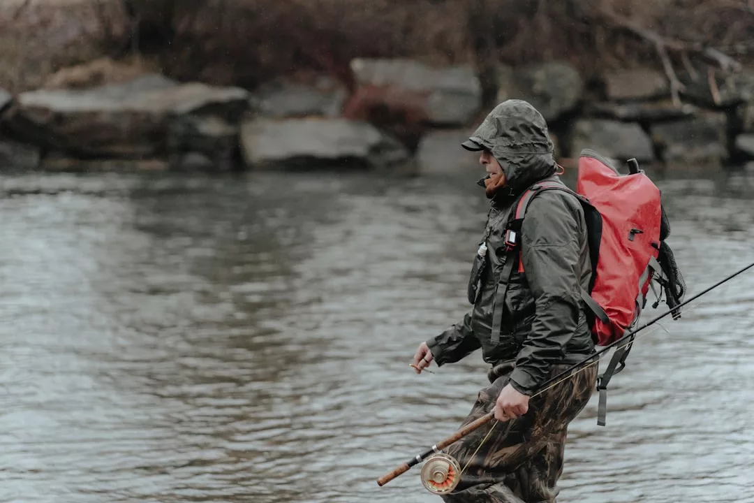 Una persona vestida con ropa de montaña y una mochila roja está de pie en un río con una caña de pescar, con la orilla rocosa de Mallorca de fondo, capturando el espíritu de aventura del lujo.