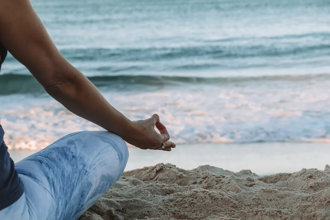 Persona disfrutando del lujo en las arenas de Mallorca, sentada con las piernas cruzadas cerca del océano y una mano en postura de meditación, mirando al agua tranquila.