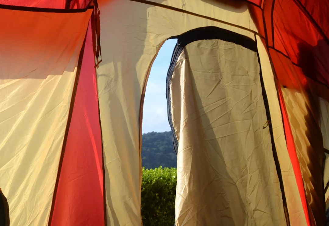 Vista desde el interior de una tienda roja y beige a través de una puerta parcialmente abierta, con la exuberante vegetación de Mallorca y una colina cubierta de árboles visible en el exterior: un toque de lujo en la naturaleza.