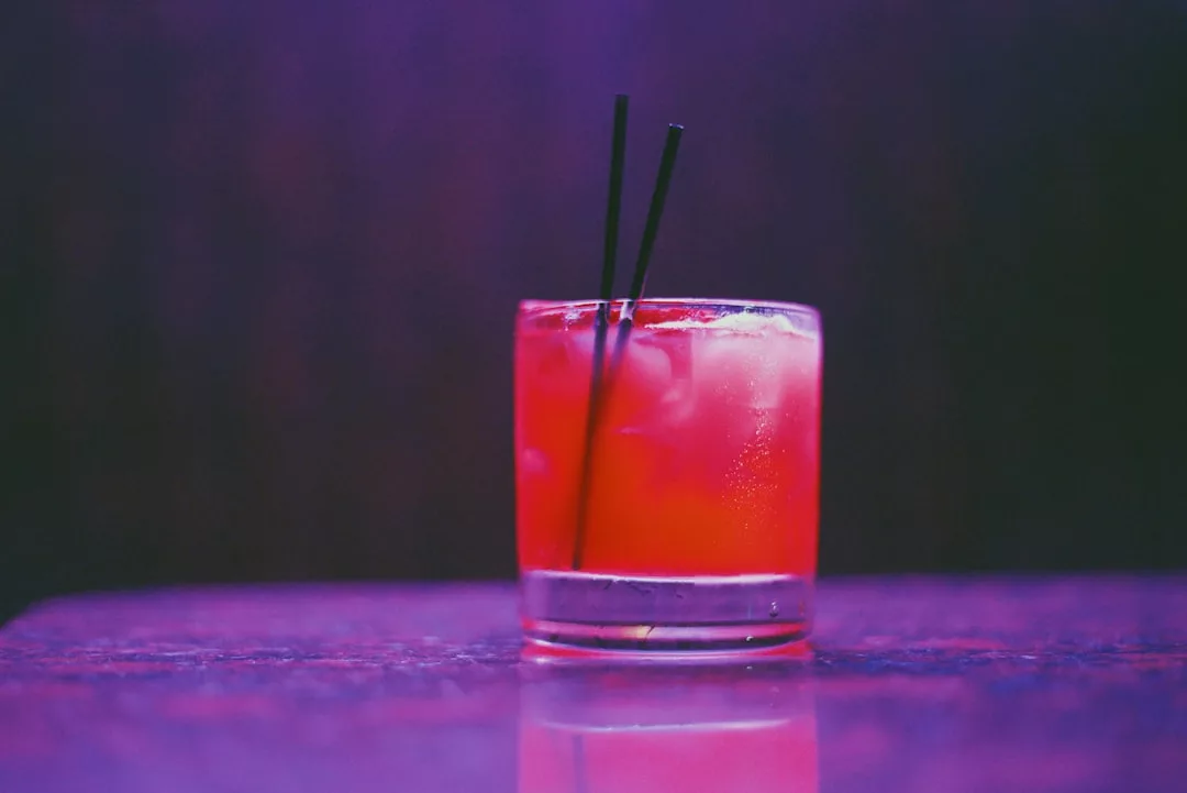 Un vaso de cóctel rojo con hielo y dos pajitas negras se asienta sobre una superficie reflectante sobre un fondo morado, capturando la esencia de Lujo en Mallorca.