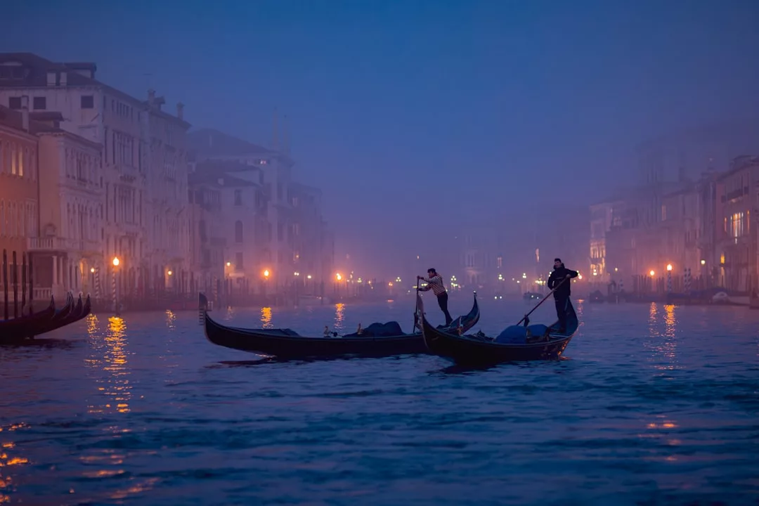 Dos góndolas con gondoleros reman a lo largo de un brumoso canal de Venecia al atardecer, la escena irradia lujo mientras los edificios históricos y las farolas brillan en el fondo, evocando recuerdos del encantador encanto de Mallorca.