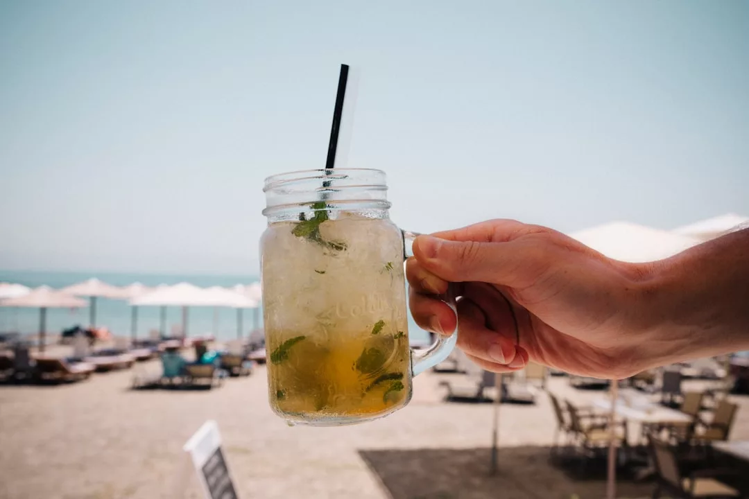 Una mano sostiene una jarra llena de una bebida de color claro y hojas de menta, con una pajita negra, disfrutando de la soleada escena de la playa de Lujo de Mallorca, con tumbonas y sombrillas al fondo.