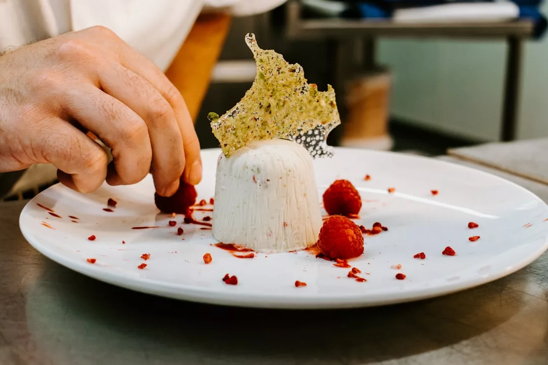 Una mano coloca una frambuesa junto a un postre emplatado coronado con un crujiente decorativo, encarnando el espíritu de Mallorca lujo, con más frambuesas y migas rojas artísticamente esparcidas por el plato blanco.