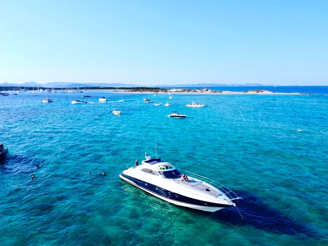 Un yate a motor blanco y azul flota en las aguas turquesas de Mallorca, rodeado de varias embarcaciones más pequeñas y una costa rocosa bajo un cielo azul despejado: una auténtica escena de lujo.