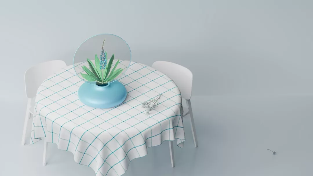 Una mesa redonda con un mantel de cuadros blancos y azules, dos sillas blancas, un jarrón azul con hojas verdes y un vasito cerca del jarrón, aportan un toque de lujo inspirado en Mallorca a un fondo de luz minimalista.