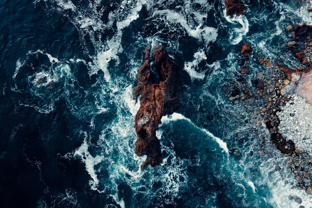 Vista aérea de las olas del mar rompiendo contra un afloramiento rocoso a lo largo de la costa de Mallorca, con rocas dispersas que añaden lujo natural a la escena.