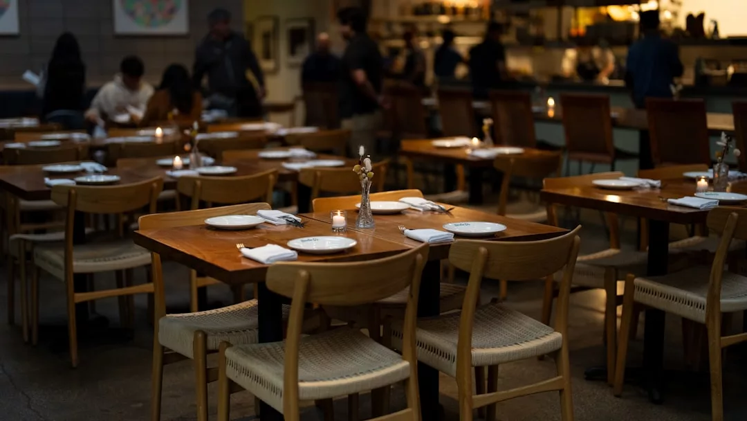 El comedor de un restaurante poco iluminado de Mallorca, con mesas y sillas de madera, platos, servilletas y velas, desprende un lujo discreto mientras unas cuantas personas disfrutan del acogedor ambiente de fondo.