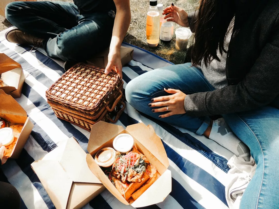Dos personas sentadas en una manta a rayas al aire libre en Mallorca, disfrutando de comida en cajas para llevar, bebidas y una cesta de picnic de mimbre para darle un toque de lujo.