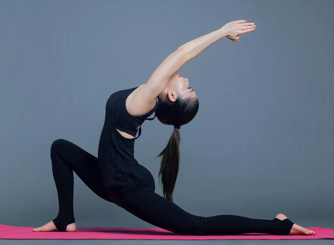 Una persona en ropa deportiva negra realiza una postura de yoga sobre una esterilla rosa, estirando una pierna hacia atrás y extendiendo los brazos hacia delante, evocando el espíritu sereno de Mallorca sobre un fondo gris liso.