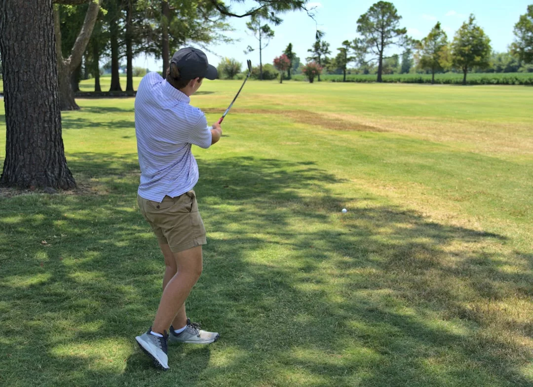 Una persona con gorra, polo y pantalón corto balancea un palo de golf en un campo de hierba de Mallorca, rodeado de árboles y manchas de luz solar, encarnando el espíritu del lujo.