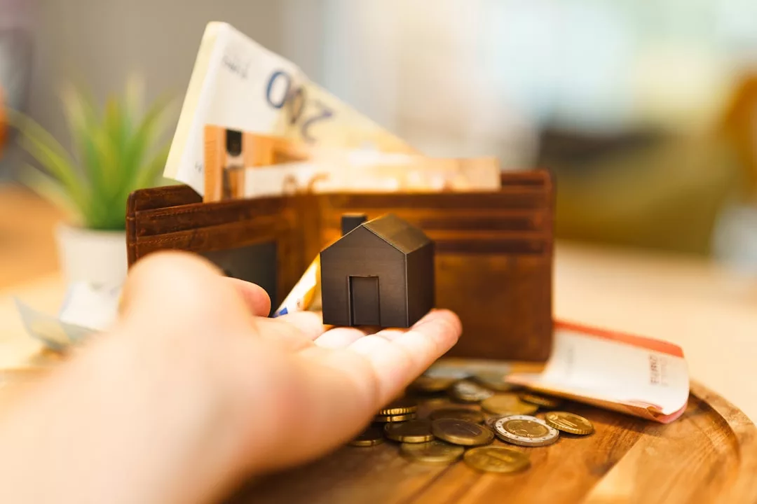 Una mano sostiene la maqueta de una pequeña casa frente a una cartera llena de billetes y monedas de euro sobre una superficie de madera, evocando el lujo de poseer una propiedad en Mallorca.