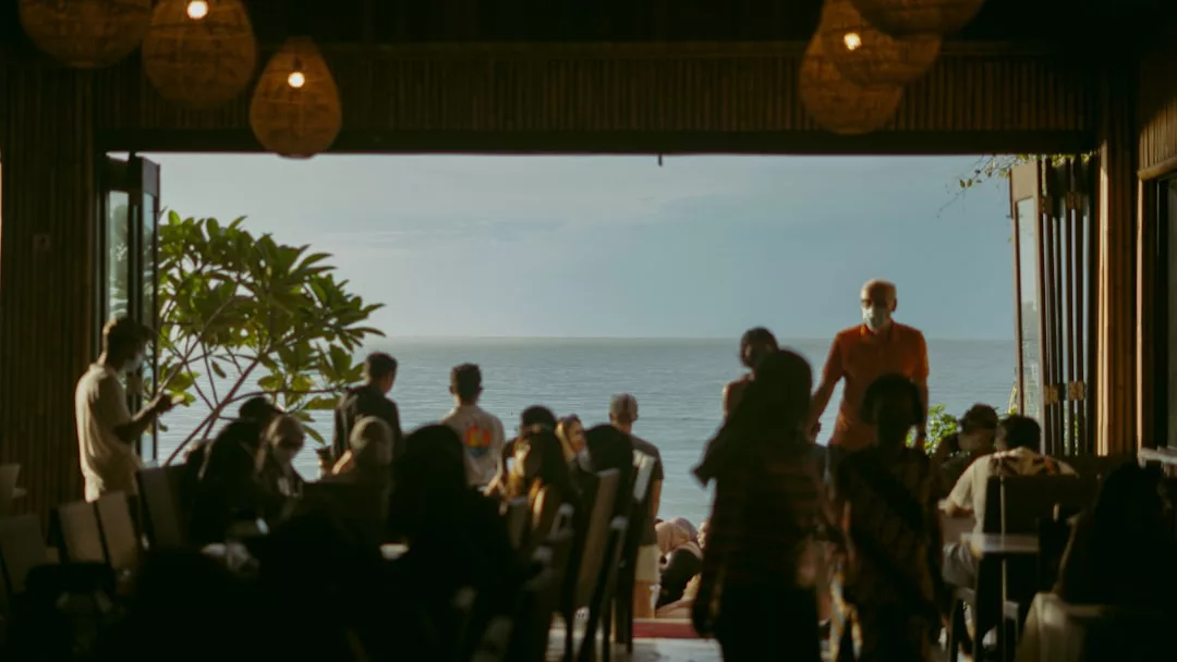 Un grupo de personas se reúnen en el interior de un restaurante costero de Mallorca tenuemente iluminado y con las puertas abiertas, mirando hacia el océano al fondo, disfrutando de una atmósfera de auténtico lujo.