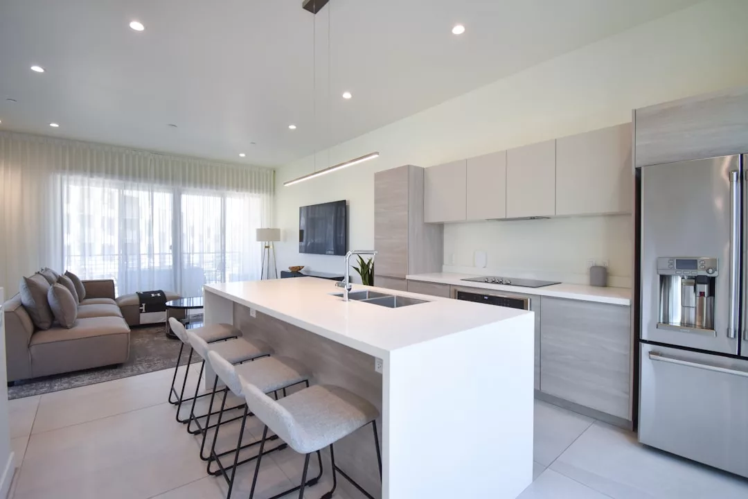 Moderna cocina con isla blanca, cuatro taburetes de bar, electrodomésticos empotrados y una zona de estar abierta con sofá, TV y una gran ventana con cortinas transparentes, perfecta para disfrutar del estilo de vida de lujo de Mallorca.