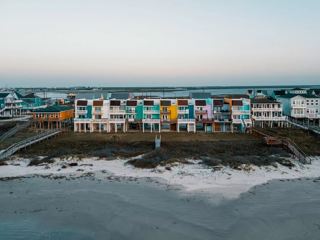 Una hilera de coloridas casas sobre pilotes frente al océano en Mallorca, exuda una sensación de lujo, con playa de arena y zona de césped en primer plano bajo un cielo despejado.