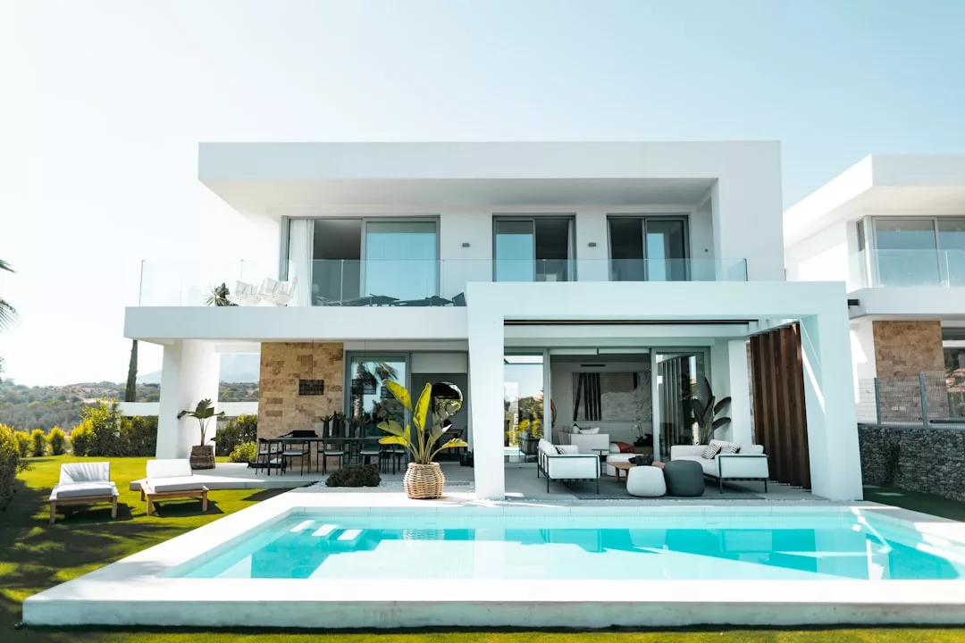 Moderna casa de dos plantas en Mallorca con grandes ventanales de cristal, un patio cubierto y una piscina rectangular rodeada de muebles de jardín y exuberante vegetación, perfecta para vivir el lujo.