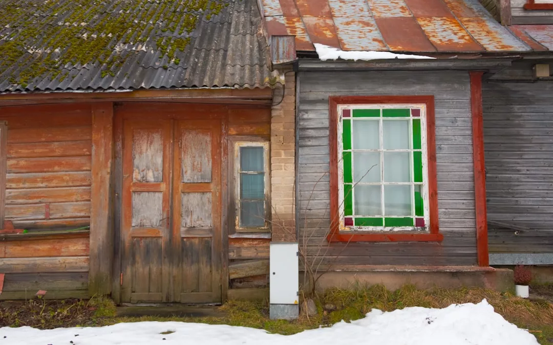 Una casa de madera desgastada con tejado de metal oxidado, musgo, pintura desconchada, puertas dobles y ventanas con bordes verdes. La nieve cubre el suelo delante, lejos del sol de Mallorca, pero con su propio lujo rústico en medio del frío.