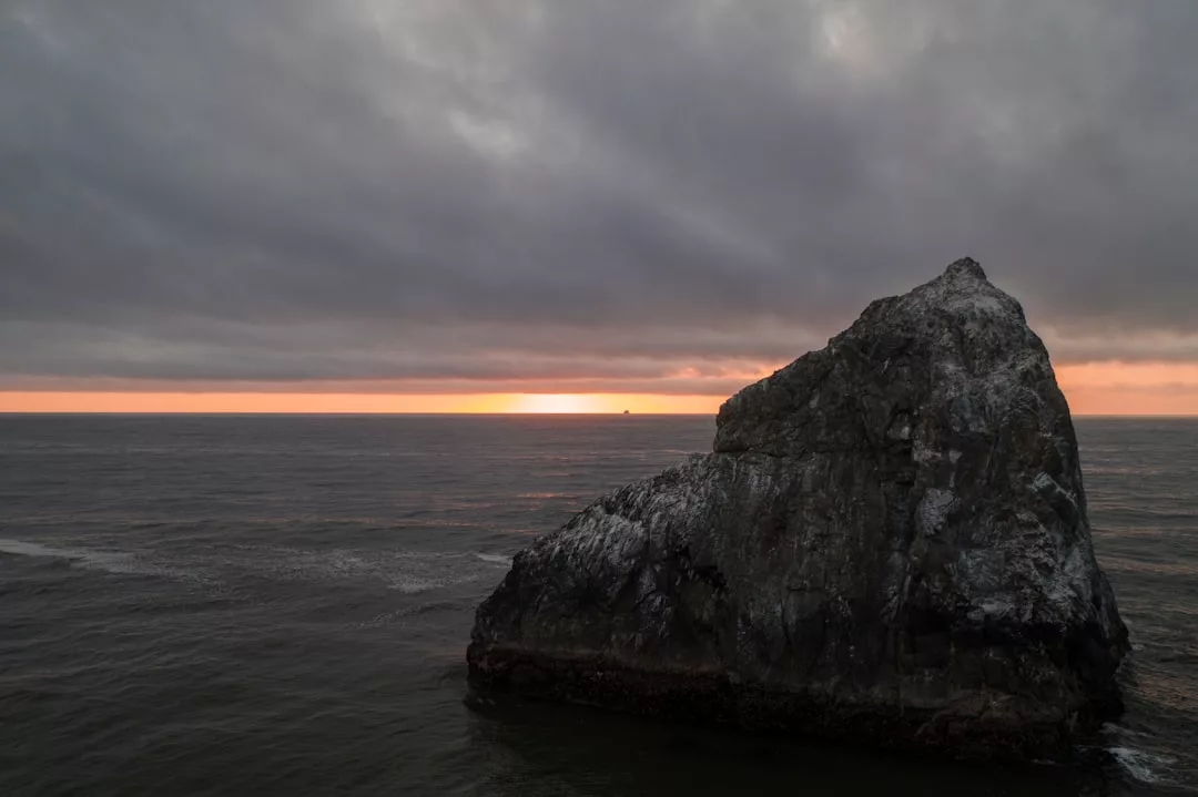 Un gran afloramiento rocoso surge del océano cerca de Mallorca, con nubes oscuras en lo alto y una estrecha banda de luz naranja en el horizonte al atardecer.