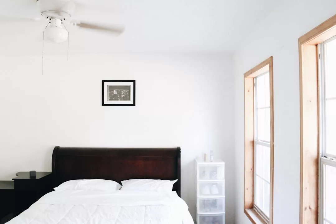 Un dormitorio minimalista en Mallorca con una cama de madera oscura, ropa de cama blanca, una mesita de noche pequeña, una cajonera de plástico blanco, dos ventanas y un ventilador de techo. Una foto enmarcada cuelga sobre la cama, añadiendo un toque de lujo a este espacio sereno.