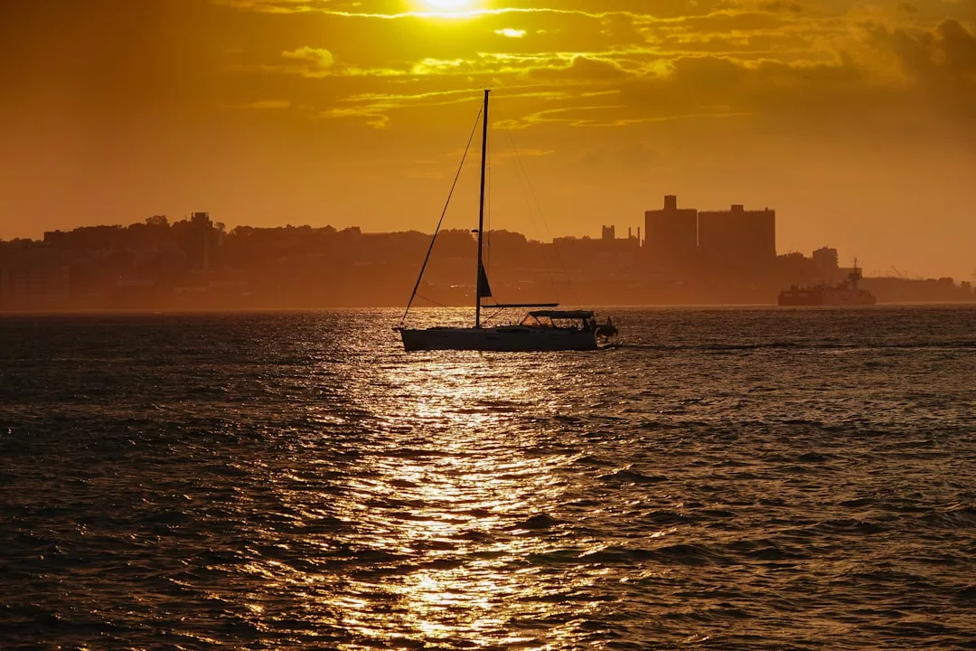 Un velero se desliza por una tranquila masa de agua al atardecer, con el sol bajo en el cielo y los edificios de la ciudad de fondo, captando la esencia del lujo y la serena belleza de Mallorca.