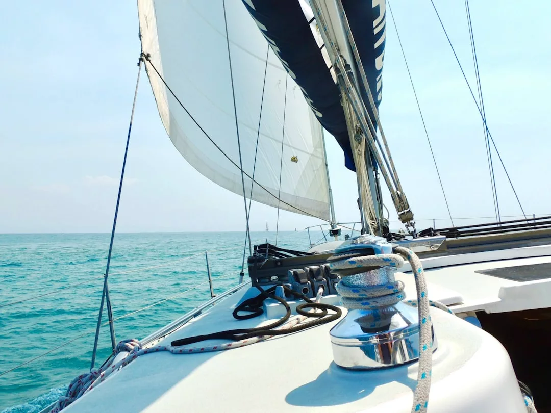 Primer plano de la cubierta de un velero con cuerdas y cabrestante en primer plano, velas blancas izadas y las tranquilas aguas azules de Mallorca bajo un cielo despejado de fondo: un ambiente de puro lujo en el mar.
