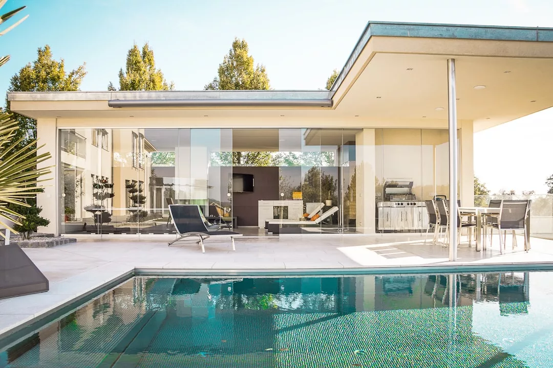 Casa moderna con paredes de cristal de suelo a techo, patio exterior, tumbonas, barbacoa, mesa de comedor y una piscina de lujo en primer plano, perfecta para disfrutar del soleado estilo de vida mallorquín.
