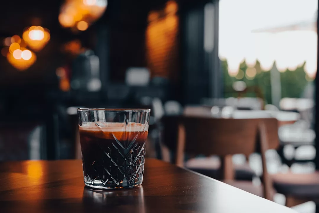 Un vaso de bebida helada oscura descansa sobre una mesa de madera en un café poco iluminado, que evoca el relajado lujo de Mallorca, con sillas desdibujadas y luces que brillan suavemente al fondo.