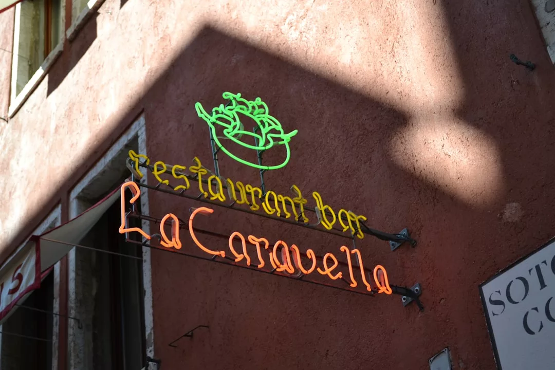 Cartel de neón que dice "restaurante bar La Caravella" en amarillo y naranja con el contorno verde de un barco, montado en una pared de estuco rojo en Mallorca, parcialmente en la sombra: un toque de lujo en cada detalle brillante.