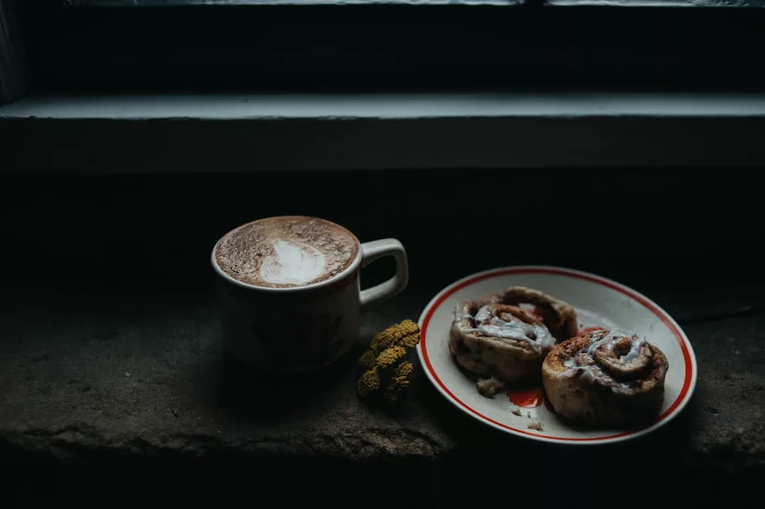 Una taza de capuchino con arte latte se sienta junto a un plato con dos rollos de canela en un oscuro alféizar, aportando un toque de lujo mallorquín junto a un pequeño racimo de flores amarillas.