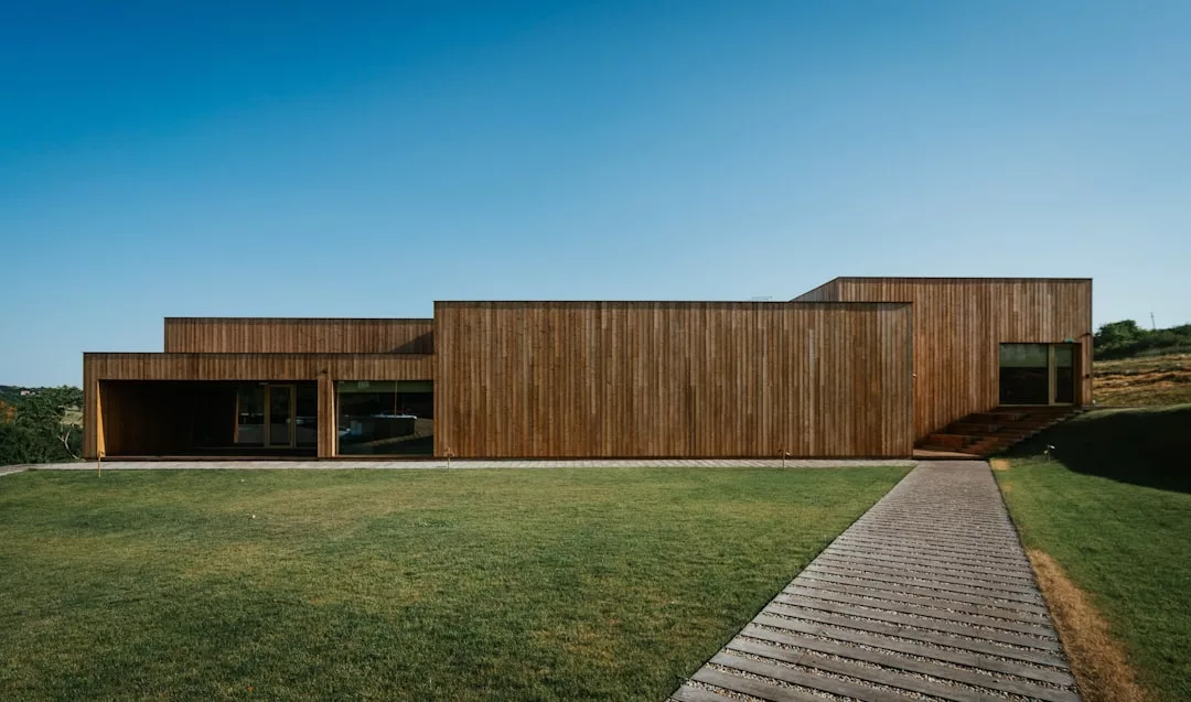 Una moderna casa rectangular de madera de lujo con grandes ventanales se asienta sobre un césped bajo el cielo azul claro de Mallorca, una pasarela de madera conduce a la entrada.
