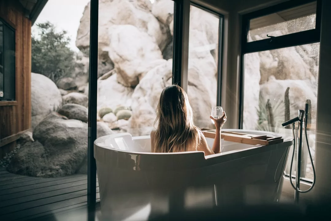 Una persona con el pelo largo está sentada en una bañera junto a grandes ventanales, sosteniendo un vaso y contemplando el paisaje rocoso de Mallorca, rodeada de un aire de lujo.