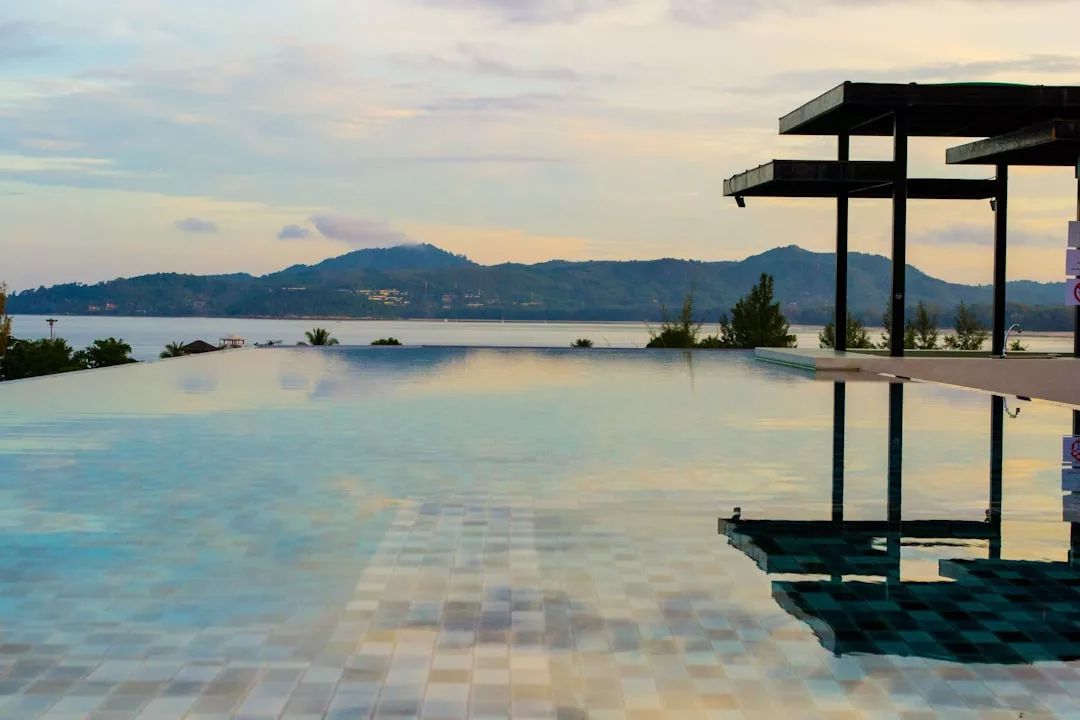 Piscina infinita con suelo de baldosas con vistas a una tranquila masa de agua y colinas lejanas en Mallorca bajo un cielo nublado, con una moderna estructura Lujo en el lado derecho.
