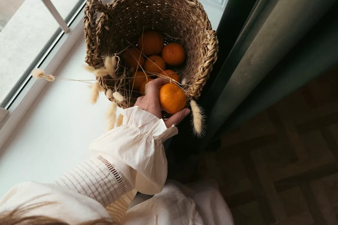 Una persona con camisa blanca sostiene una naranja en una cesta tejida llena de naranjas y hierba seca, capturando el relajado lujo de Mallorca cerca de una ventana iluminada por el sol.