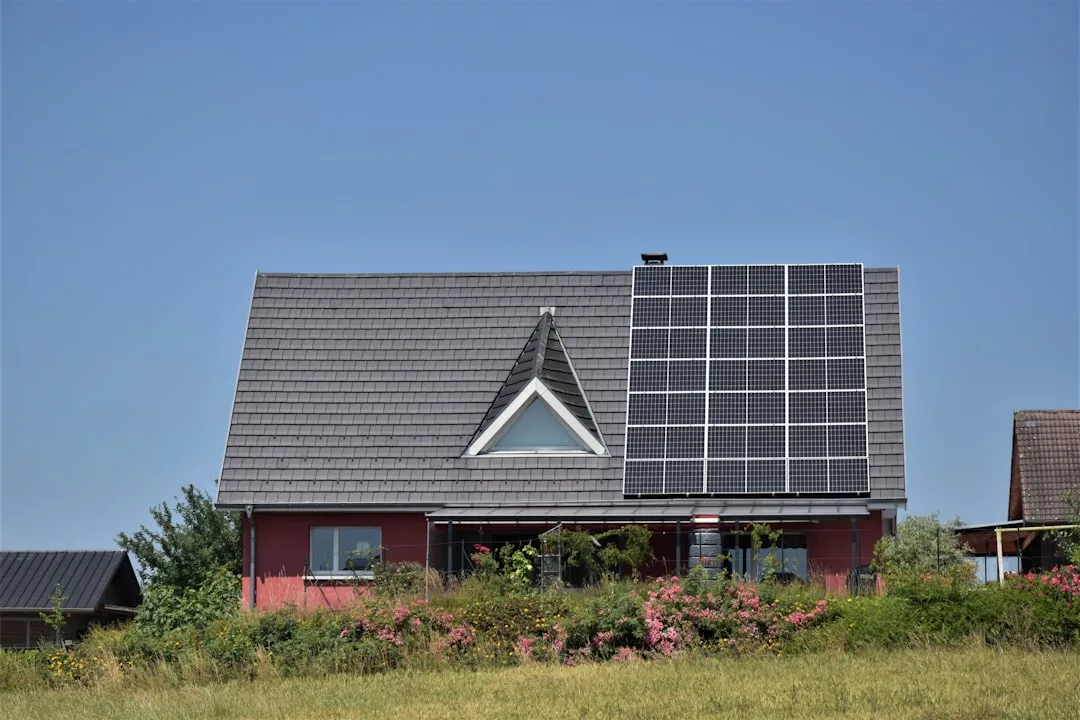 Una casa con un empinado tejado gris cuenta con paneles solares en un lateral; flores rosas y exuberante vegetación llenan el patio delantero bajo un cielo azul despejado, capturando la esencia de la vida de lujo en la hermosa Mallorca.