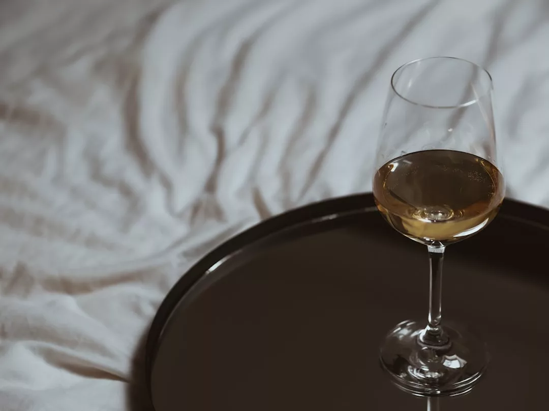 Una copa de vino blanco reposa en una bandeja oscura, colocada sobre una colcha blanca desaliñada: un acogedor toque de Lujo inspirado en la relajada elegancia de Mallorca.