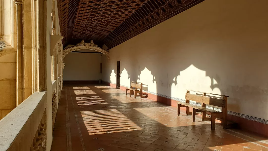 Un pasillo soleado en Mallorca con bancos de madera, suelo de baldosas, un decorativo techo arqueado e intrincadas sombras proyectadas en la pared desde las ventanas ornamentadas: un espacio acogedor que destila lujo a cada paso.