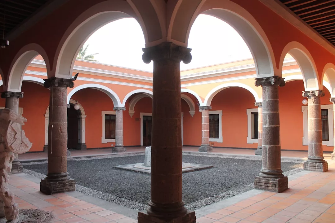 Vista de un patio de estilo colonial en Mallorca, con columnas de piedra, arcos y una zona central abierta rodeada de paredes naranjas y ventanas que evocan una sensación de lujo.