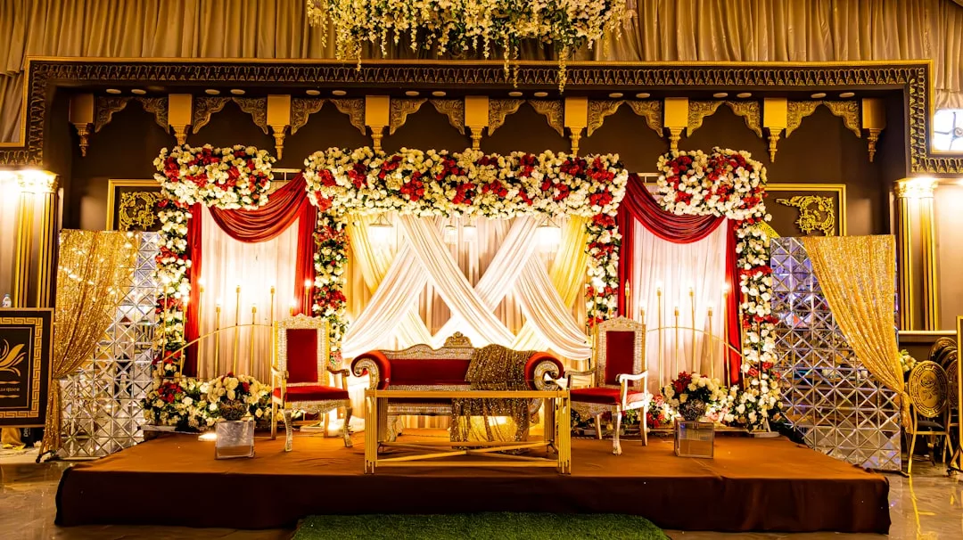 Un escenario de boda decorado con flores rojas y blancas, telas drapeadas, un sofá central, dos sillas y una cálida iluminación captan la esencia del lujo, perfecto para una celebración inolvidable en Mallorca.