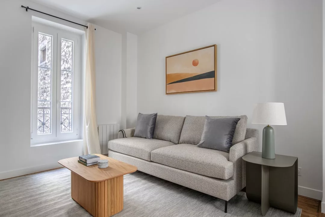Un moderno salón con un sofá gris claro, dos cojines grises, una mesa de centro de madera y arte abstracto enmarcado en una pared blanca, que destila lujo inspirado en la serena elegancia de Mallorca.