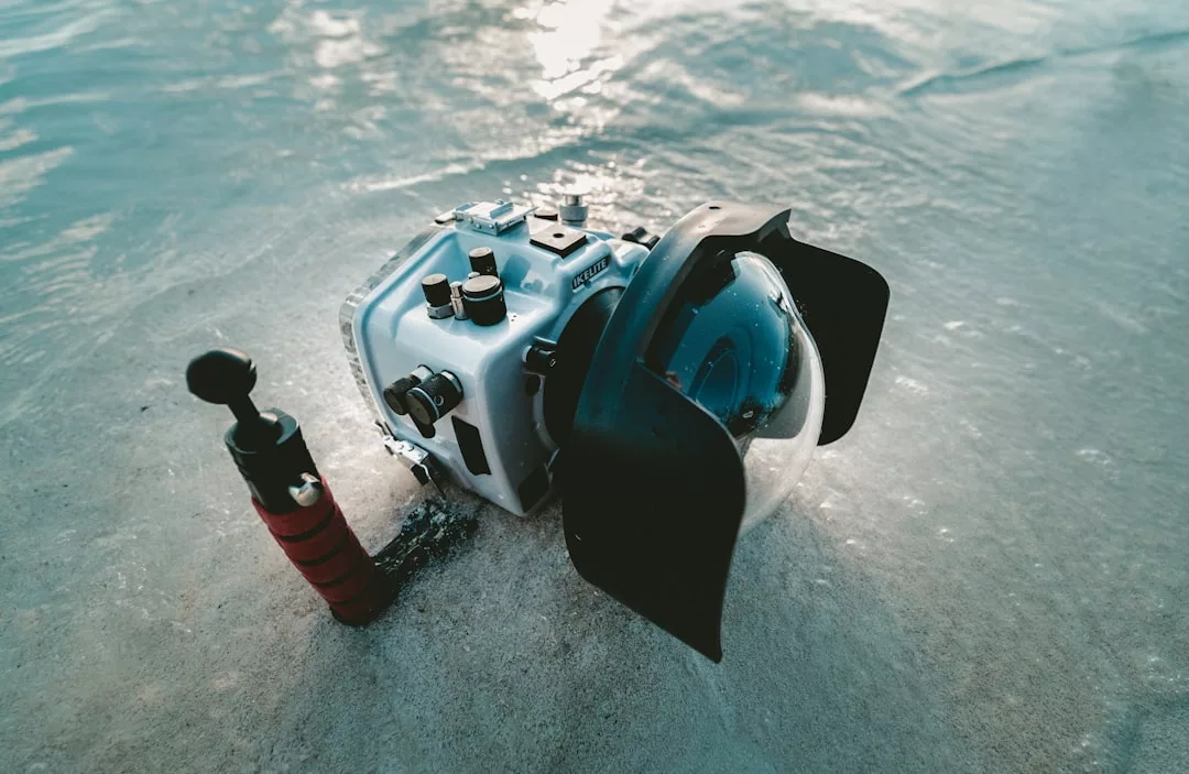 Una carcasa de cámara subacuática Lujo y un pequeño accesorio de agarre descansan sobre la arena mojada de la costa de Mallorca, con el suave chapoteo del agua a su alrededor.