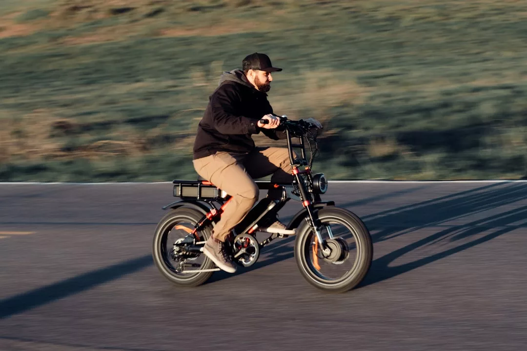 Una persona con gorra y chaqueta conduce una bicicleta eléctrica por una carretera asfaltada de Mallorca, con la hierba difuminada al fondo, captando un momento de lujo y estilo.